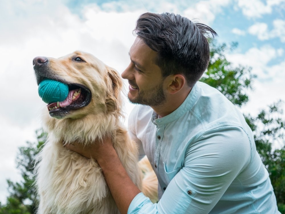 A man smiles while holding a golden retriever that has a teal ball in its mouth, with trees and a cloudy sky in the background.